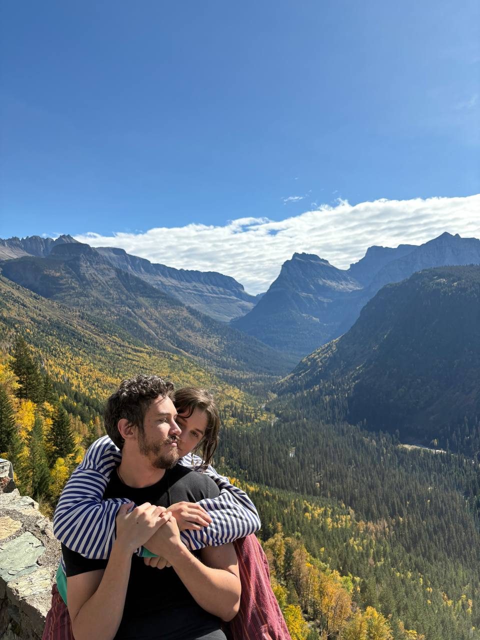 Therese hugging Thomas from behind above a forested valley